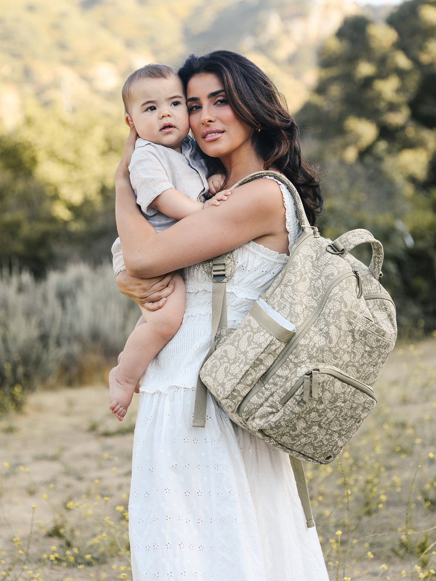A mother holding her baby, wearing the JuJuBe Classic Diaper Backpack in Paisley Floral